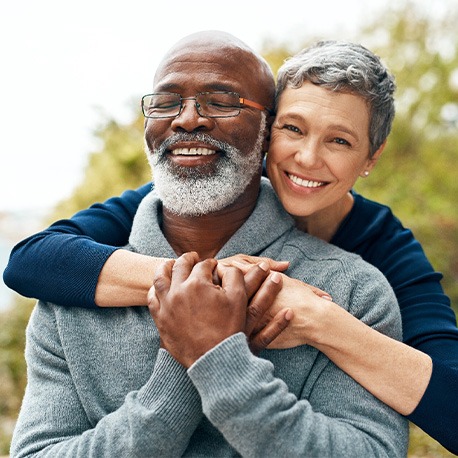 Woman in blue shirt hugging man in gray sweater from behind outside smiling